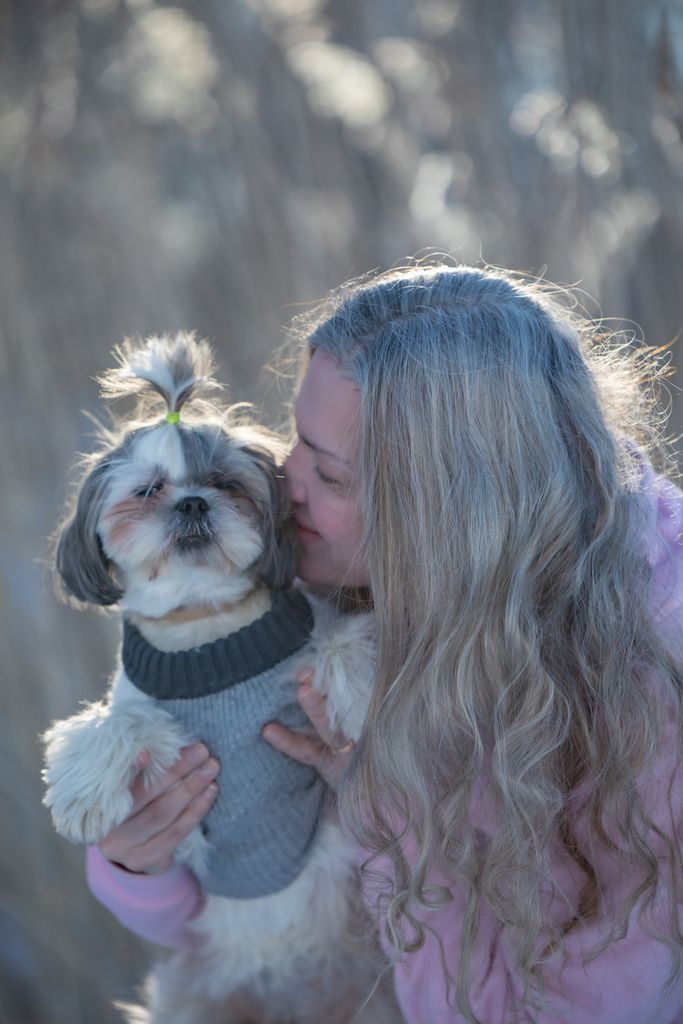 Caucasian woman with long blonde hair hugging her small, fluffy dog outdoors in bright daylight.